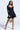 A woman stands confidently in the IN THE MOOD BANDAGE MINI DRESS, a fitted long-sleeve black mini with a flared hem. She holds a leopard print handbag, wears black strappy heels, and has long wavy hair. The background is plain light gray.