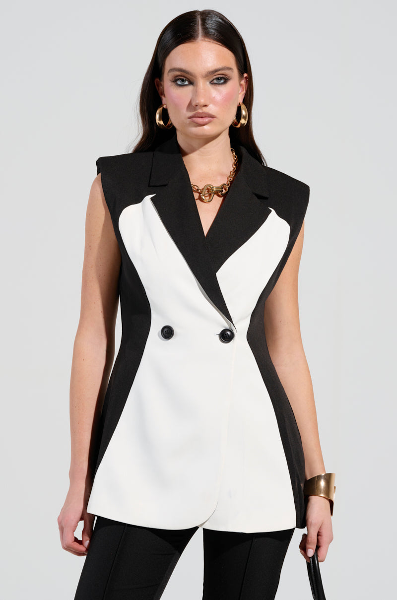 A confident woman poses in the LABOR OF LOVE COLOR BLOCK BLAZER VEST, highlighting its sleeveless silhouette and structured design alongside chic black pants. She accessorizes with gold hoop earrings, a chunky necklace, and a wide bracelet. Her long, dark hair is styled straight.