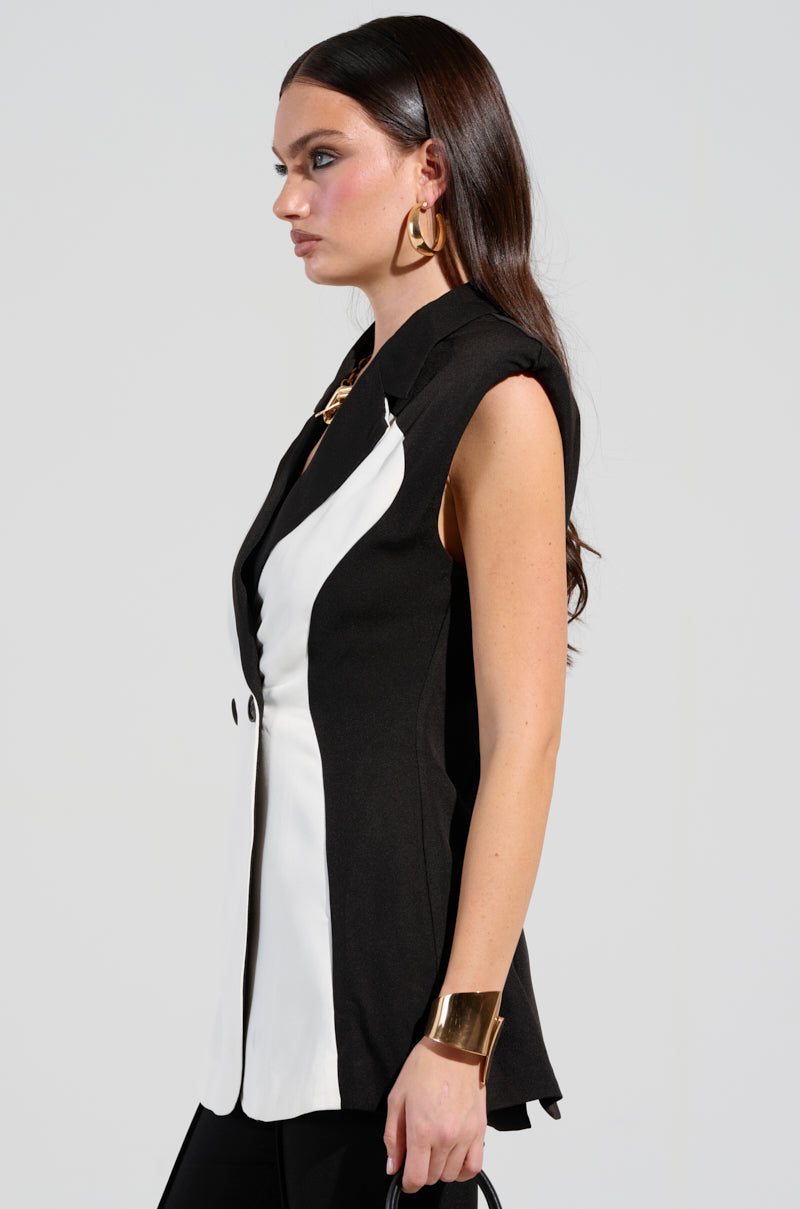 A woman with long dark hair models the LABOR OF LOVE COLOR BLOCK BLAZER VEST, showcasing it as a structured piece with a deep V-neck. She complements her outfit with large gold hoop earrings and a gold cuff bracelet. The sleeveless silhouette is highlighted in profile view against a plain background.