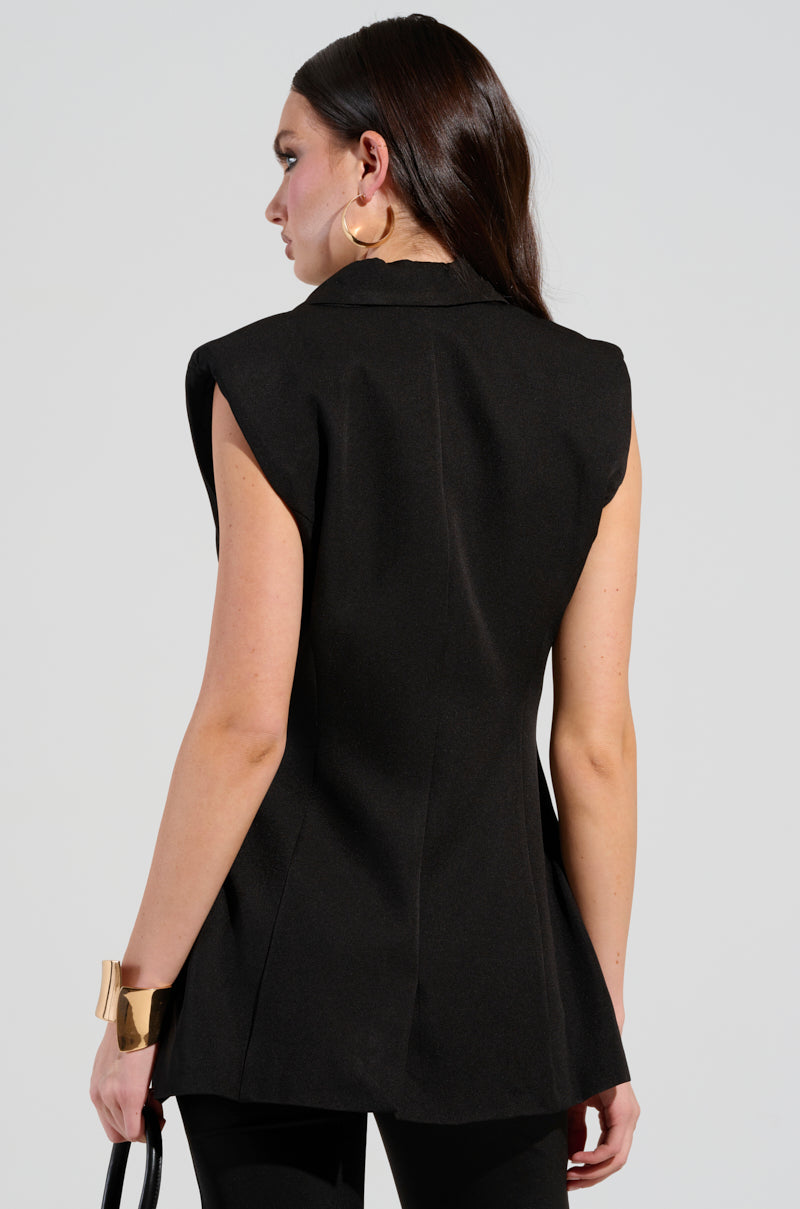 A woman with long, dark hair stands sideways, facing away. She wears a sleek sleeveless silhouette featuring the LABOR OF LOVE COLOR BLOCK BLAZER VEST and matching pants, accessorized with large gold hoop earrings and a wide gold bracelet. In her hand is a black handbag against the plain light gray background.