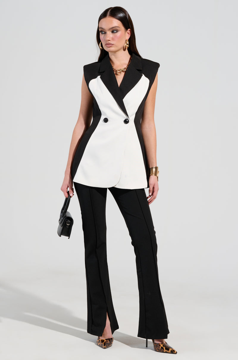 A woman is elegantly standing in the LABOR OF LOVE COLOR BLOCK BLAZER VEST, showcasing its stylish sleeveless silhouette. She pairs it with sleek black pants and holds a small black purse while wearing leopard print heels. Her ensemble is complemented by gold necklace and bracelet accessories, all framed against a plain background.