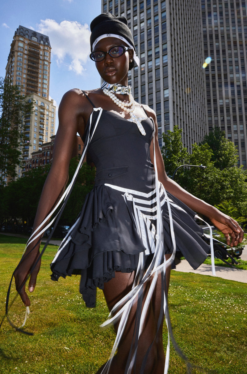 Confidently posing outdoors in a city park among tall buildings, a stylish person wears the SPORTY SPICE RUFFLE MINI DRESS with white ribbons, paired with a headwrap, glasses, layered necklaces, and sunlight accentuating their look.