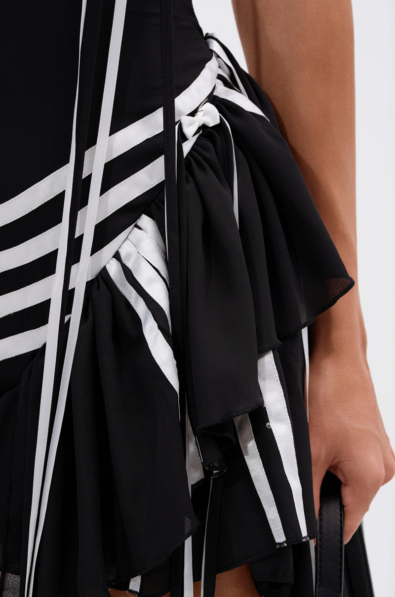 Close-up of the SPORTY SPICE RUFFLE MINI DRESS, an athletic-chic LBD featuring white diagonal stripes and ruffle layers. Shows a partial arm and hand holding a small black bag; the fabric is flowy and detailed.