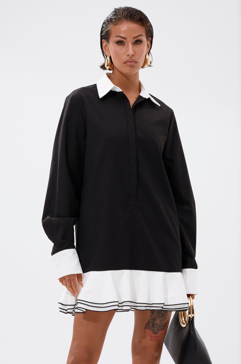 A woman exudes confidence in the ALWAYS CLASSY POPLIN SHIRT DRESS, featuring a black design with white cuffs, collar, and ruffled hem. She accessorizes with a black bag, gold hoop earrings, and wet-look hair against a plain backdrop.