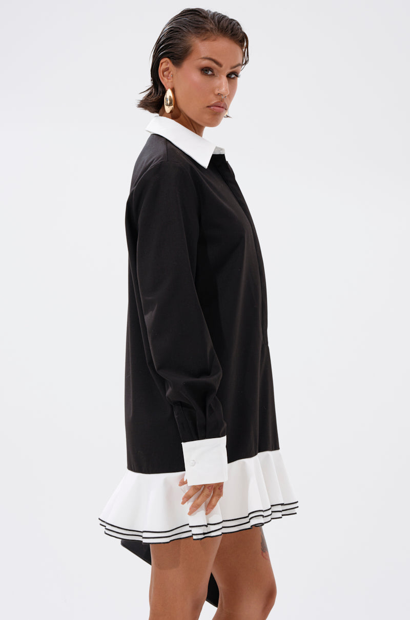 A woman models the ALWAYS CLASSY POPLIN SHIRT DRESS, a black mini dress with white cuffs, a white collar, and a ruffled skirt accented by black stripes, posing sideways against a plain white background.
