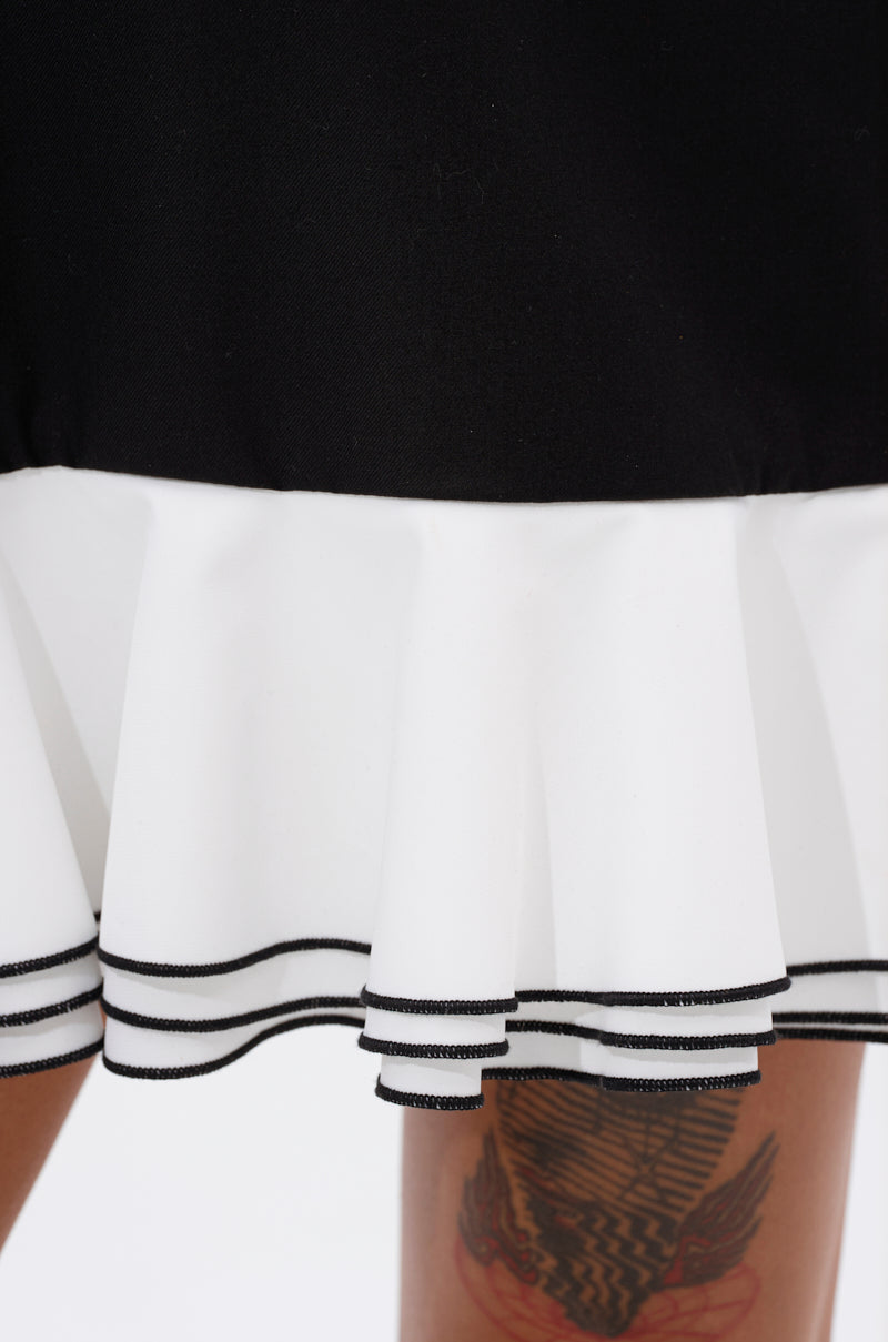 Close-up of the hem of the ALWAYS CLASSY POPLIN SHIRT DRESS, featuring black and white ruffles with black trim and a glimpse of a tattooed leg beneath.