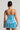 A woman is standing with her back to the camera, exuding Cinderella vibes in the ALL ABOUT ME SATIN BUBBLE MINI DRESS, a strapless, metallic blue mini dress featuring a ruched, textured design. She holds a small silver clutch in her right hand and has her hair styled in a neat, sleek bun, completing her princess-inspired look.