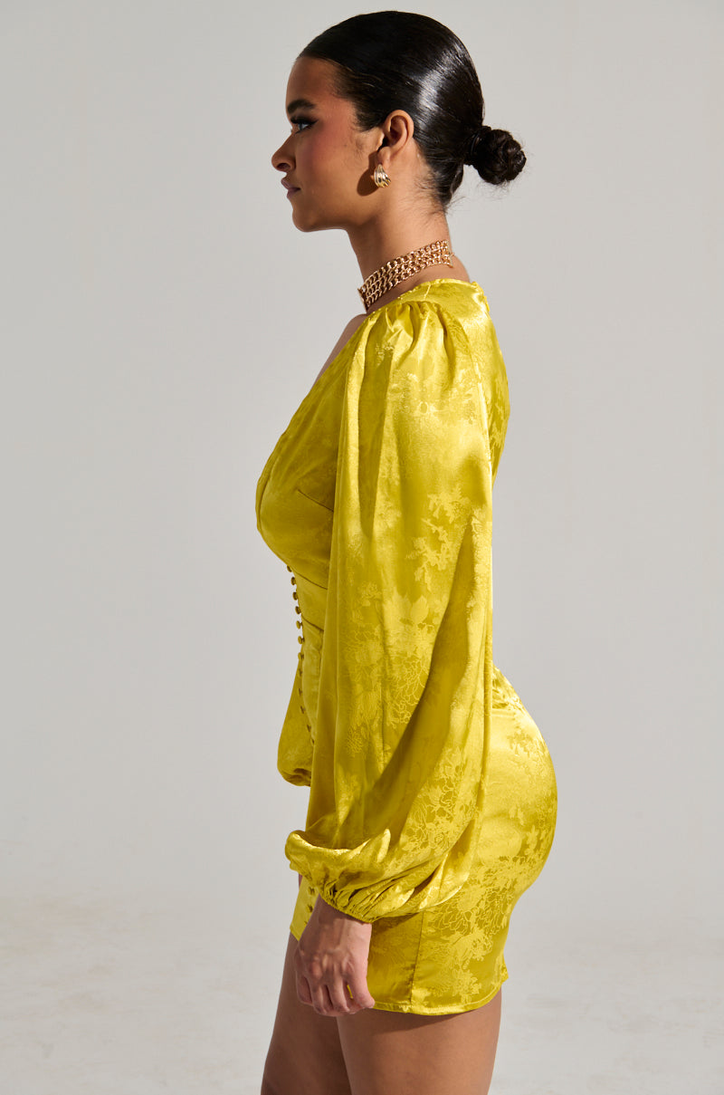 A woman in profile radiates elegance in the SLEEK AND CHIC SATIN MINI DRESS, featuring long puffed sleeves and button detailing. Her hair is styled in a sleek bun, complemented by gold jewelry including earrings and a necklace. The scene is set against a neutral background. 3