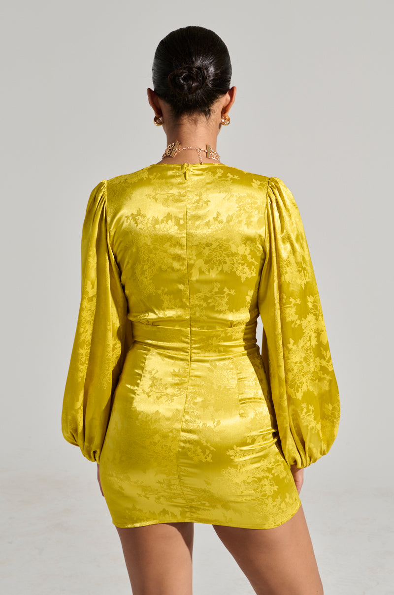 Rear view of a person wearing an elegant ensemble: the "Sleek and Chic Satin Mini Dress," a bright yellow, long-sleeved satin mini dress adorned with a floral pattern. The fitted dress boasts a short hemline, and the person has their hair tied up, accessorized with a gold necklace. 4