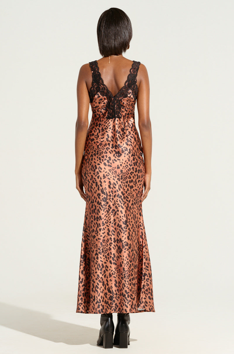 A person with short hair wears the "LEOPARD PRINT IS THE NEW BLACK SATIN MIDI DRESS," a sleeveless, ankle-length creation featuring black lace trim on the back. As they stand facing away against a plain background, the dress flows elegantly to their ankles.
