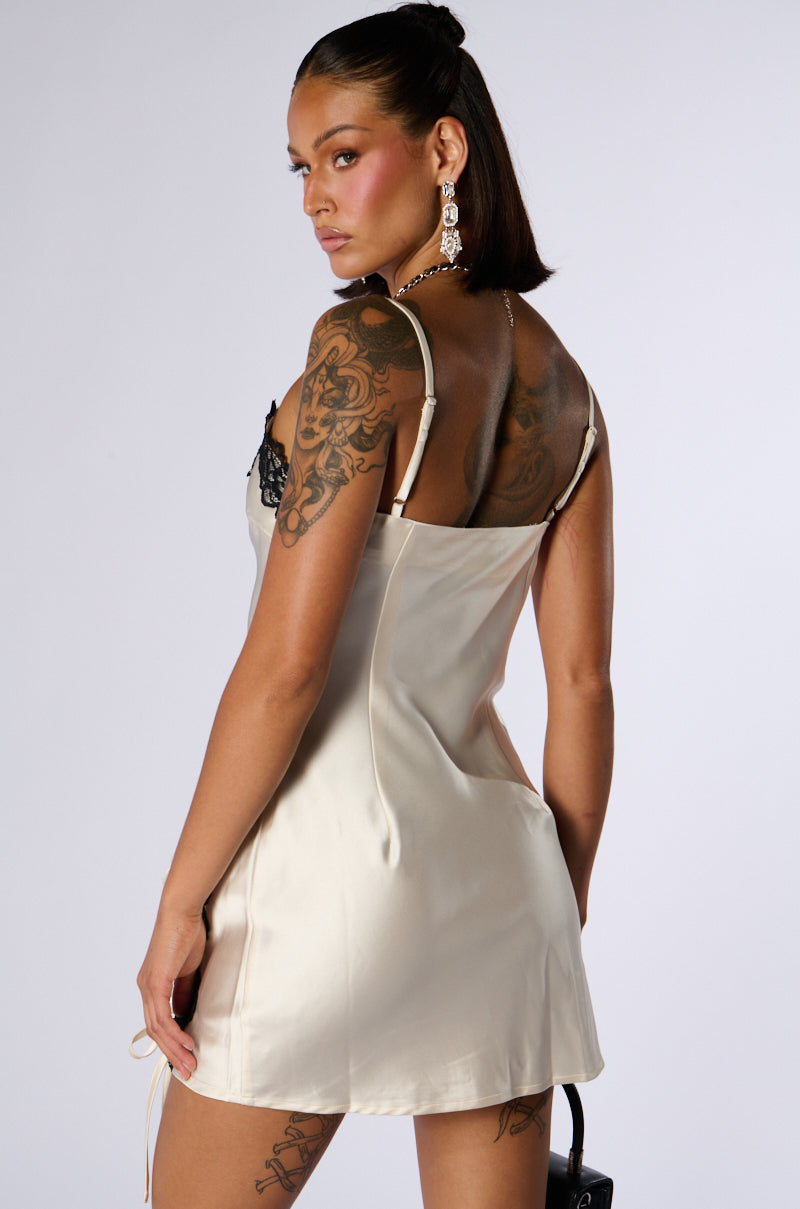 A woman with tattoos on her left arm and shoulder stands with her back to the camera, wearing the BONNIE SATIN AND LACE MINI DRESS. The dress is white satin adorned with black lace detailing and features a sweetheart neckline. She has long, dark hair pulled back and is accessorized with statement earrings. She looks over her shoulder with a neutral expression.