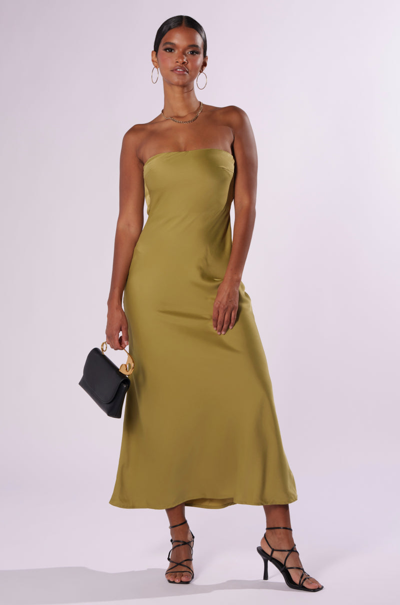 A woman stands against a plain background, wearing the BEST GUEST SATIN MAXI DRESS in olive green. The strapless neckline of the dress highlights her curve-hugging silhouette. She accessorizes with a small black handbag featuring a gold ring handle, complemented by strappy black heels and large hoop earrings.