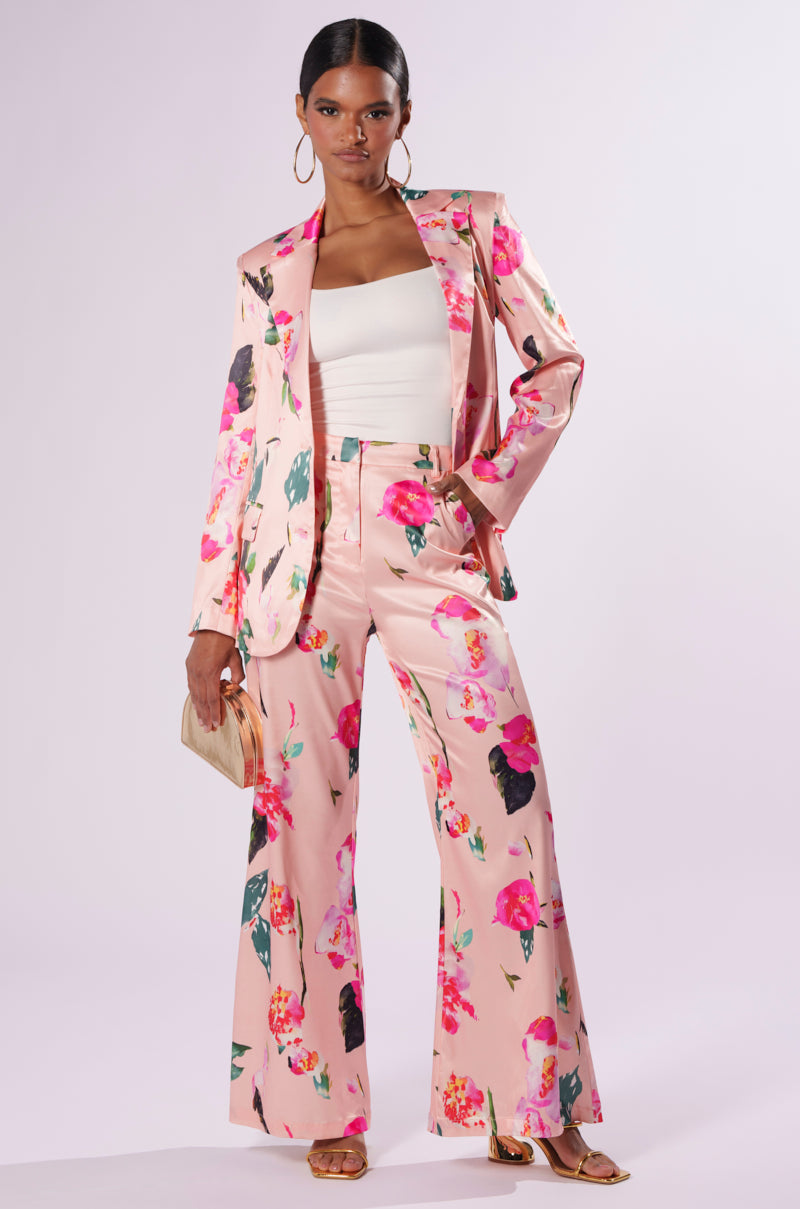 A person models a fashionable outfit featuring a pink floral blazer and matching BLOSSOM SATIN TROUSERS with a chic wide-leg silhouette. They wear a white strapless top underneath, paired with hoop earrings, holding a beige clutch, and wearing strappy sandals. The background is neutral.