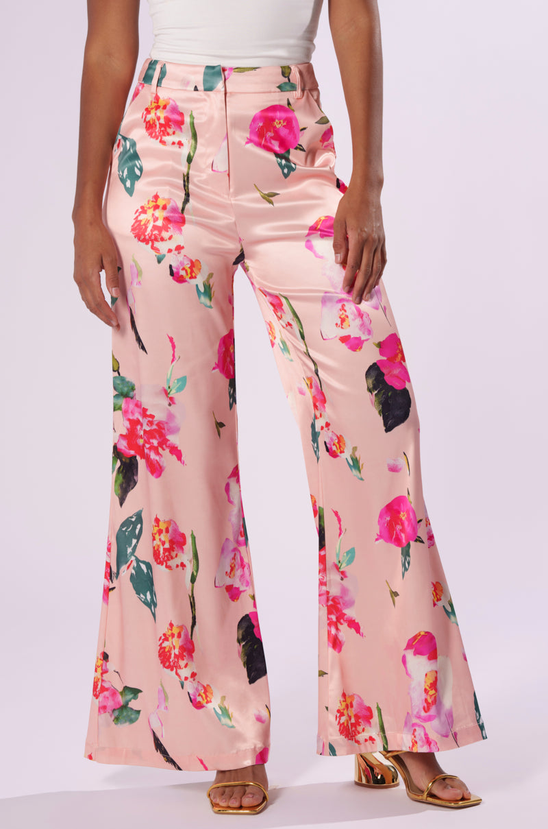 A person dressed in BLOSSOM SATIN TROUSERS adorned with a pink floral pattern. The wide-leg silhouette showcases vibrant flowers in shades of pink and green leaves. Complementing this, the person is wearing a white top tucked into the waistband and gold open-toe heels.