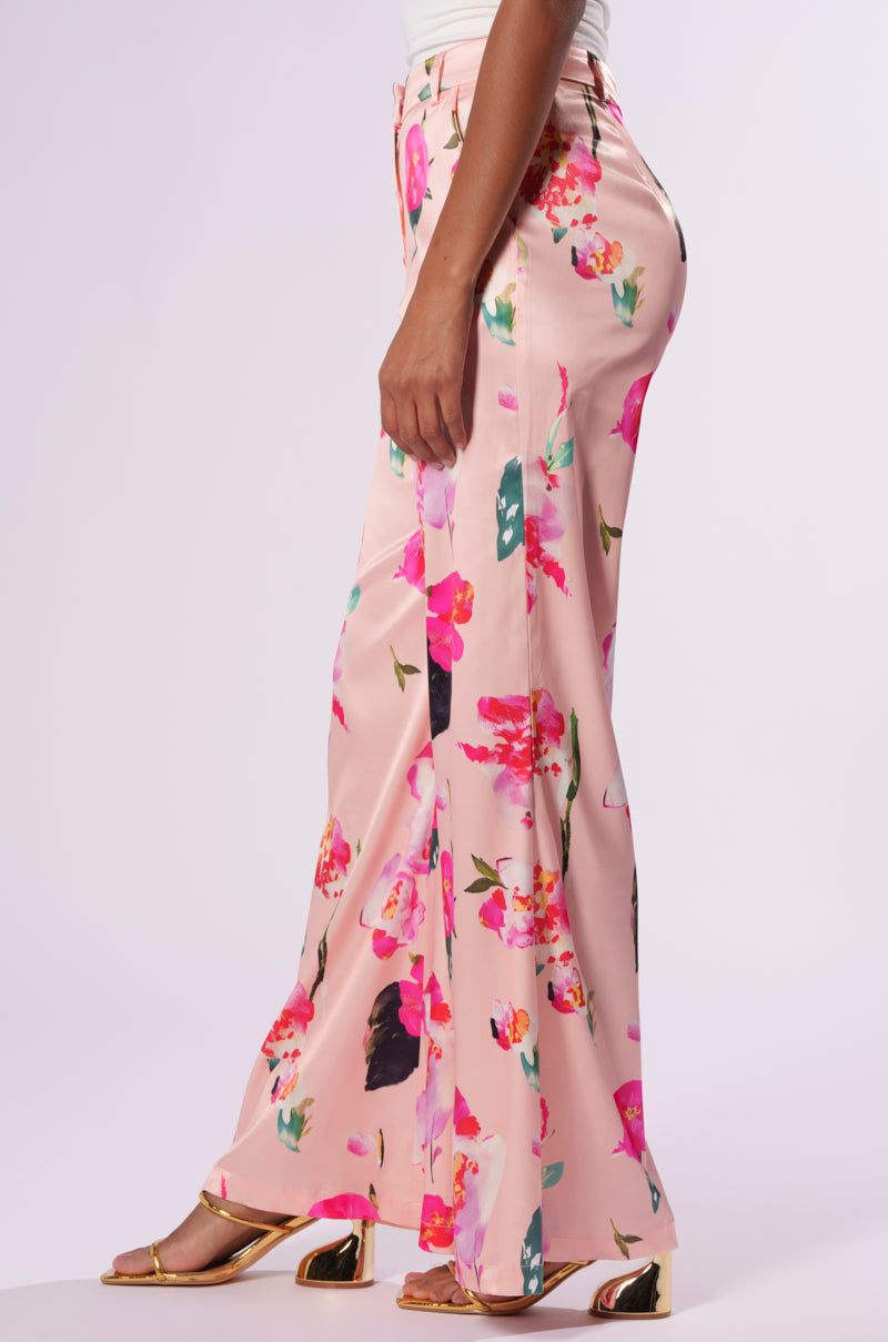 A person models the BLOSSOM SATIN TROUSERS, which feature a wide-leg silhouette and a vibrant floral pattern in shades of pink, red, and green. They pair it with a white top and gold open-toed shoes with a slight heel, set against a plain white background.