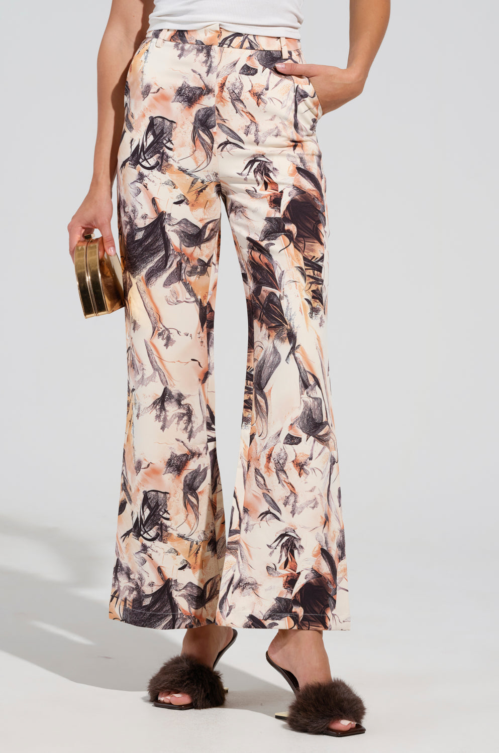 A person wearing the IANA HIGH RISE TROUSER with a vibrant floral print, paired with a white top and holding a gold clutch. They are also sporting dark fluffy high-heeled sandals. The plain light background enhances the chic yet playful essence of this business casual outfit.
