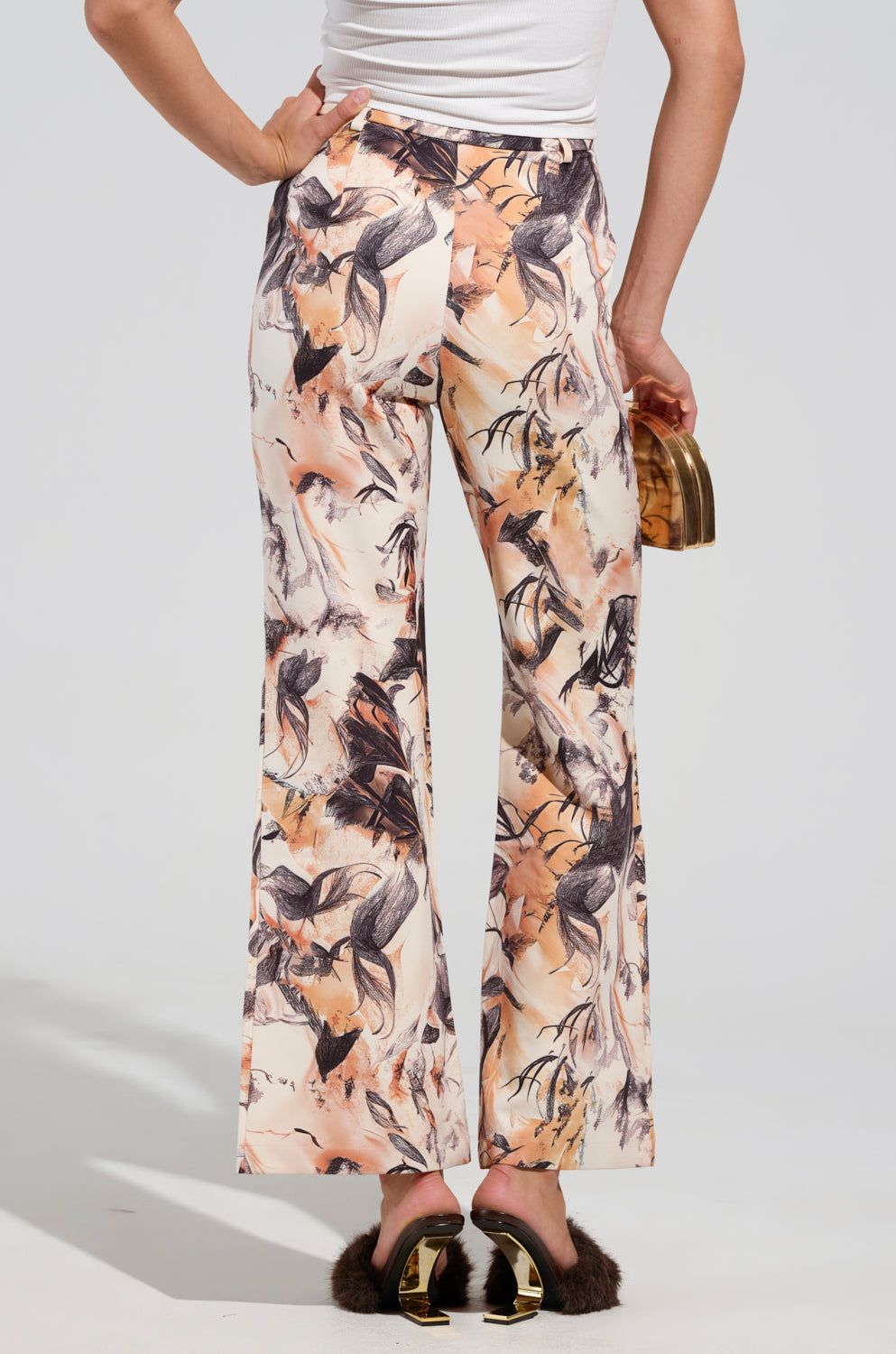 Dressed in the stylish IANA HIGH RISE TROUSER with an abstract floral design, this individual showcases a chic business casual look. They complement their outfit with a small, shiny gold clutch and black fur-lined slide sandals, all set against a simple light gray backdrop.