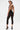 A person models the SABRINA MESH PRINTED JUMPSUIT, featuring a sheer mesh fabric, crisscross back, and cropped legs that reveal a detailed back tattoo. The look is styled with black high heels and hoop earrings.