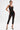A woman models the SABRINA MESH PRINTED JUMPSUIT—sleeveless, cropped at the ankles, and sheer black mesh over black undergarments. She pairs it with black gold-tipped heels and has short styled hair, set against a plain white background.
