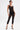 A woman exudes confidence in the SABRINA MESH PRINTED JUMPSUIT, layered over black undergarments with black and gold pointed-toe heels. Short hair and gold earrings add bold style against a plain white background.