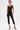 A woman with short hair and visible tattoos poses against a plain white background, confidently wearing the SABRINA MESH PRINTED JUMPSUIT over black underwear and gold-toed black heels.