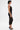 A woman in profile wears the SABRINA MESH PRINTED JUMPSUIT—an elegant sleeveless piece in sheer black lace—paired with black heels featuring gold accents, gold earrings, and a geometric upper arm tattoo for bold style.