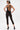 A person with short two-toned hair and visible tattoos poses facing away, showcasing the SABRINA MESH PRINTED JUMPSUIT with crisscross straps and high heels against a plain white background, highlighting bold statement style.