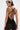 With short, highlighted hair and visible tattoos, they wear the SABRINA MESH PRINTED JUMPSUIT—a sheer, black crisscross-back style—showcasing bold statement fashion as they turn slightly away from the camera.