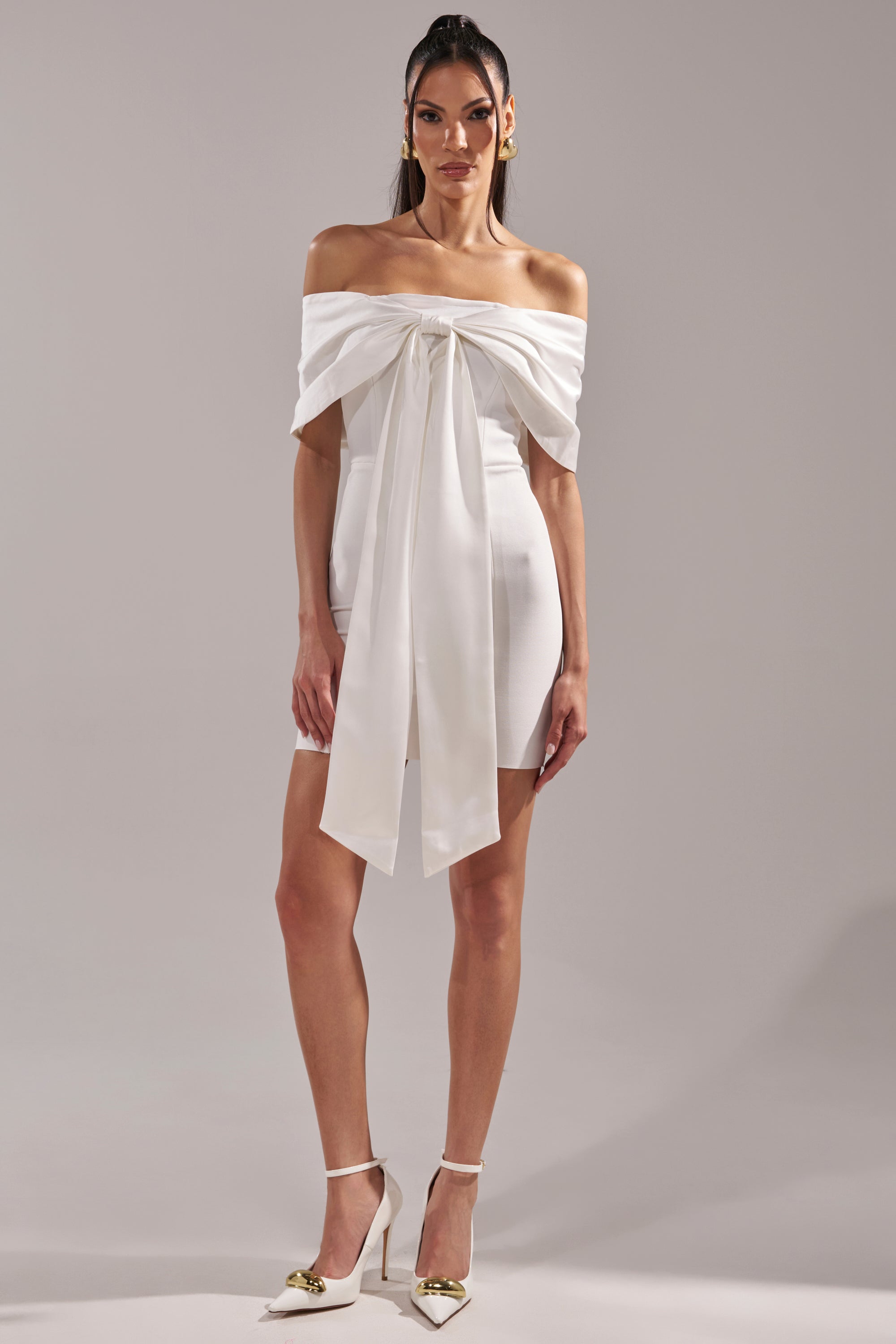 A woman faces forward in the BEEN THE GIFT SATIN BOW DRESS—an off-the-shoulder mini dress—paired with matching white high heels with gold tips, set against a plain gray background.