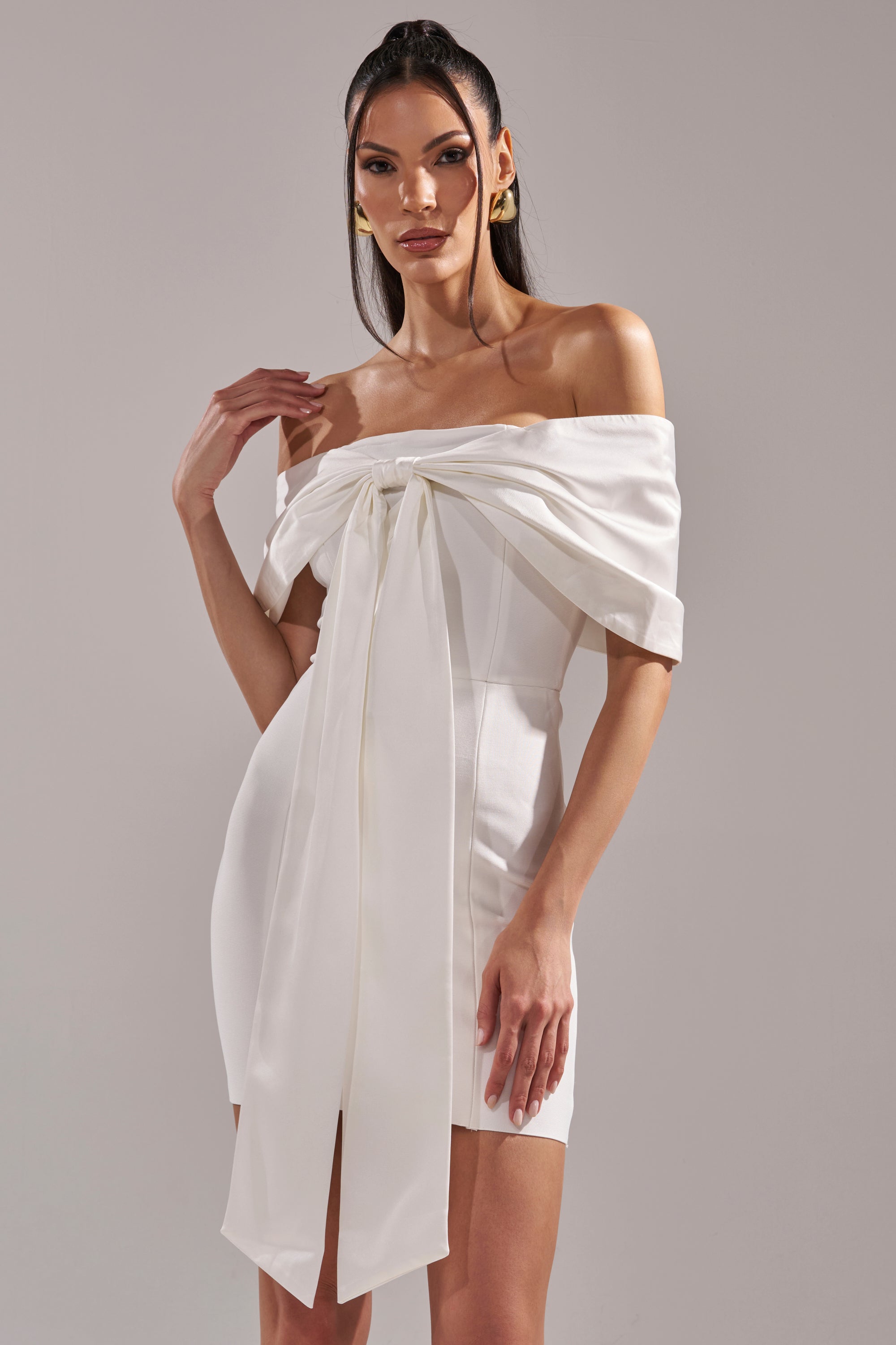 A woman models the BEEN THE GIFT SATIN BOW DRESS—a white off-the-shoulder dress featuring a large front bow and long satin ribbon—against a plain light gray background.