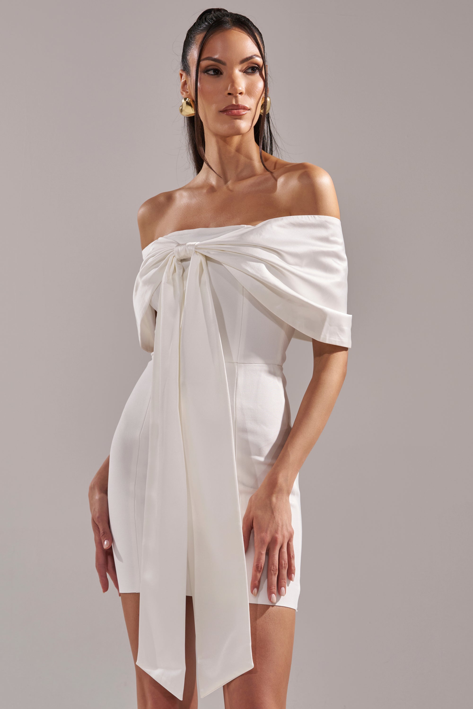 A woman models the BEEN THE GIFT SATIN BOW DRESS—an off-the-shoulder, fitted white satin dress with a large front bow and long ribbon detail—against a gray background. She has long dark hair, gold earrings, and a confident look.