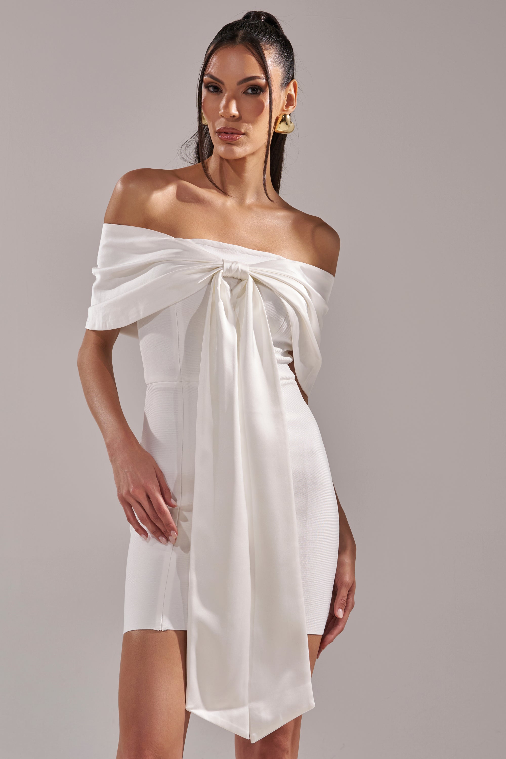 A woman models the BEEN THE GIFT SATIN BOW DRESS, featuring an off-the-shoulder neckline and white satin fabric, as she stands confidently against a plain background.