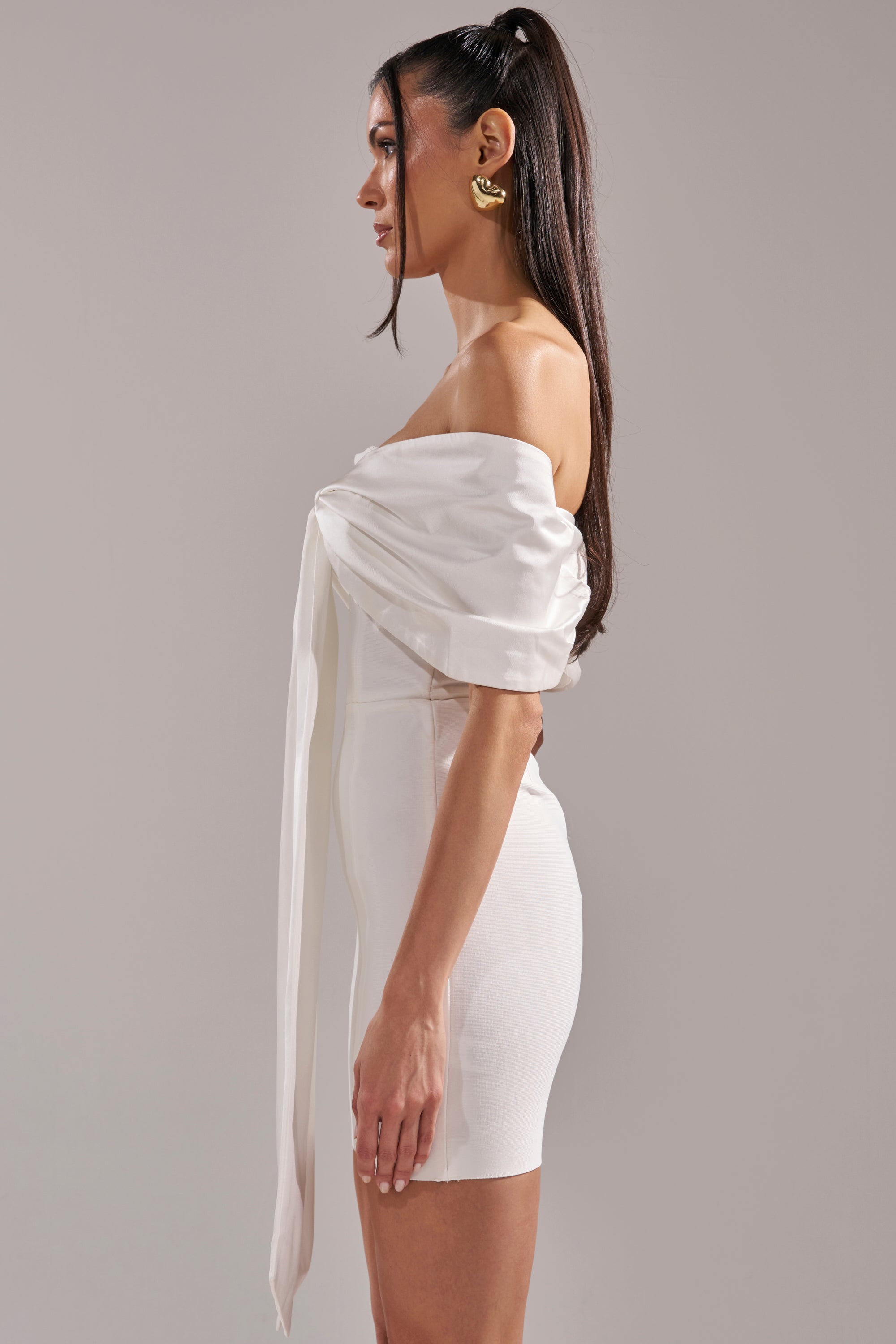 A woman stands in profile against a light gray background, wearing the BEEN THE GIFT SATIN BOW DRESS—an off-the-shoulder white formal mini dress with draped satin bow detail—paired with gold hoop earrings.