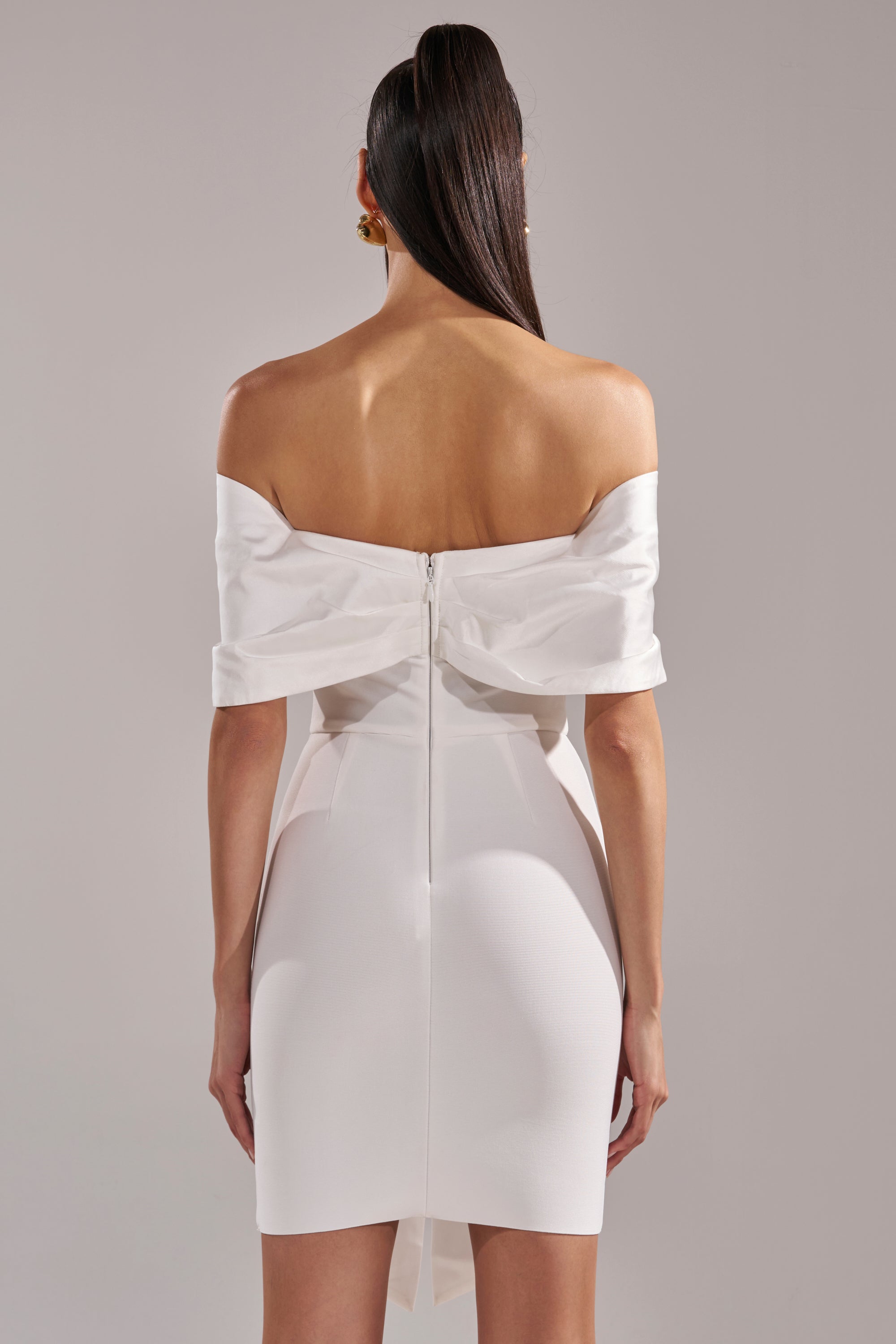 A woman with long, straight hair stands against a plain background, shown from behind, wearing the BEEN THE GIFT SATIN BOW DRESS—an off-the-shoulder white mini dress with a visible back zipper.
