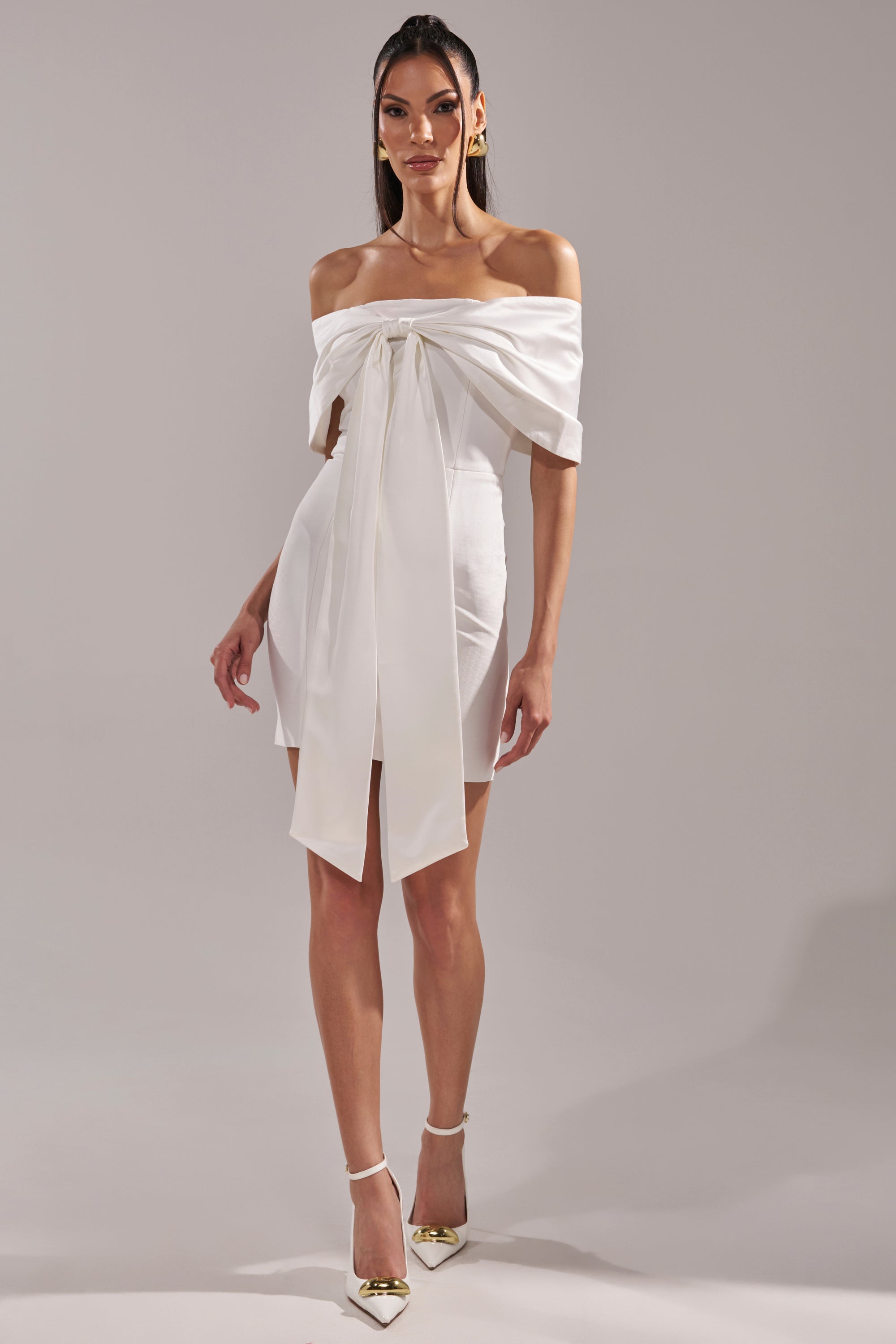 A woman poses against a neutral backdrop in the BEEN THE GIFT SATIN BOW DRESS, an off-the-shoulder style with a large front tie. She finishes her formal look with white pointed-toe heels accented by gold tips.