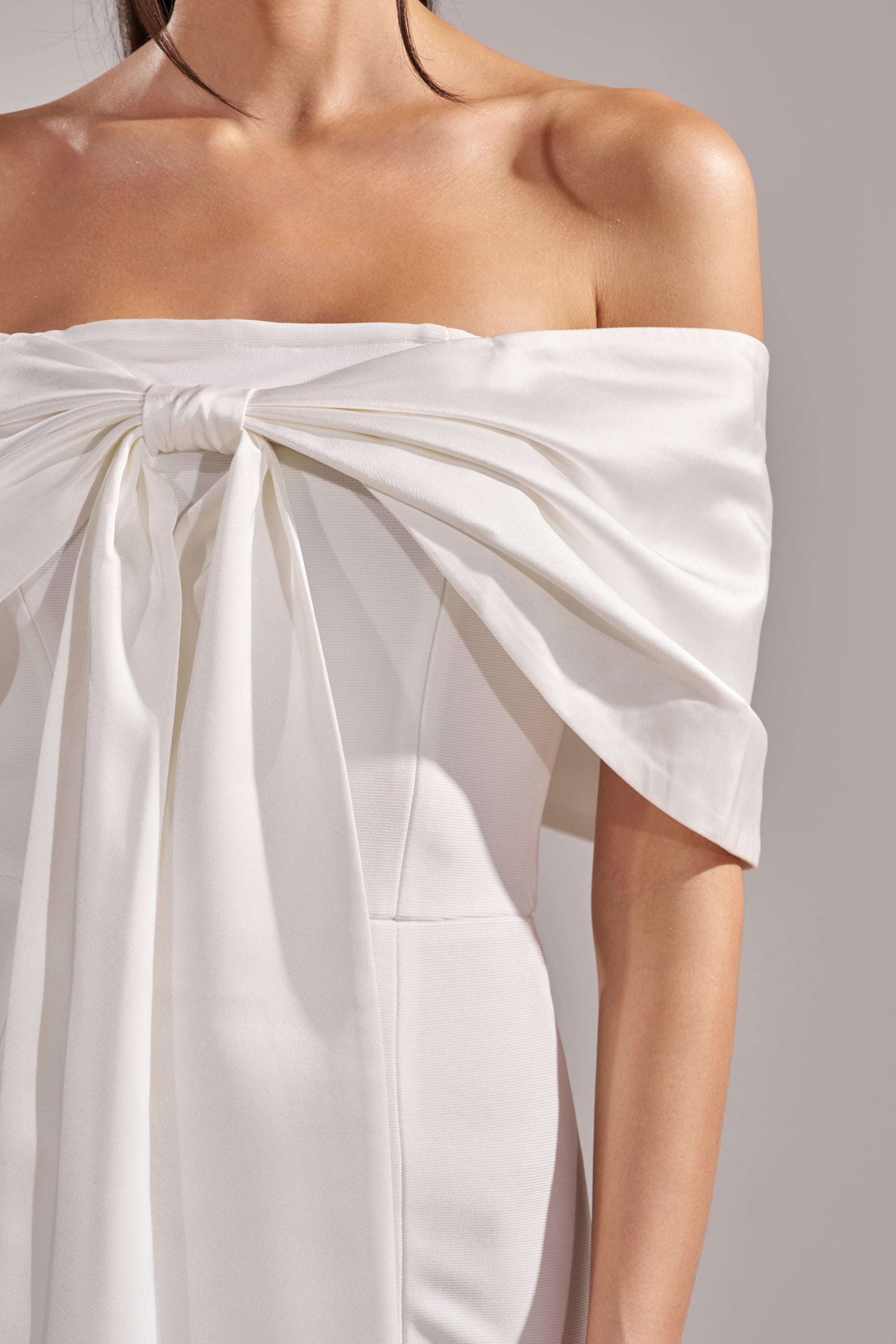 A woman models the BEEN THE GIFT SATIN BOW DRESS, featuring an off-the-shoulder white satin design with a large chest bow, shown from neck to waist against a neutral background.