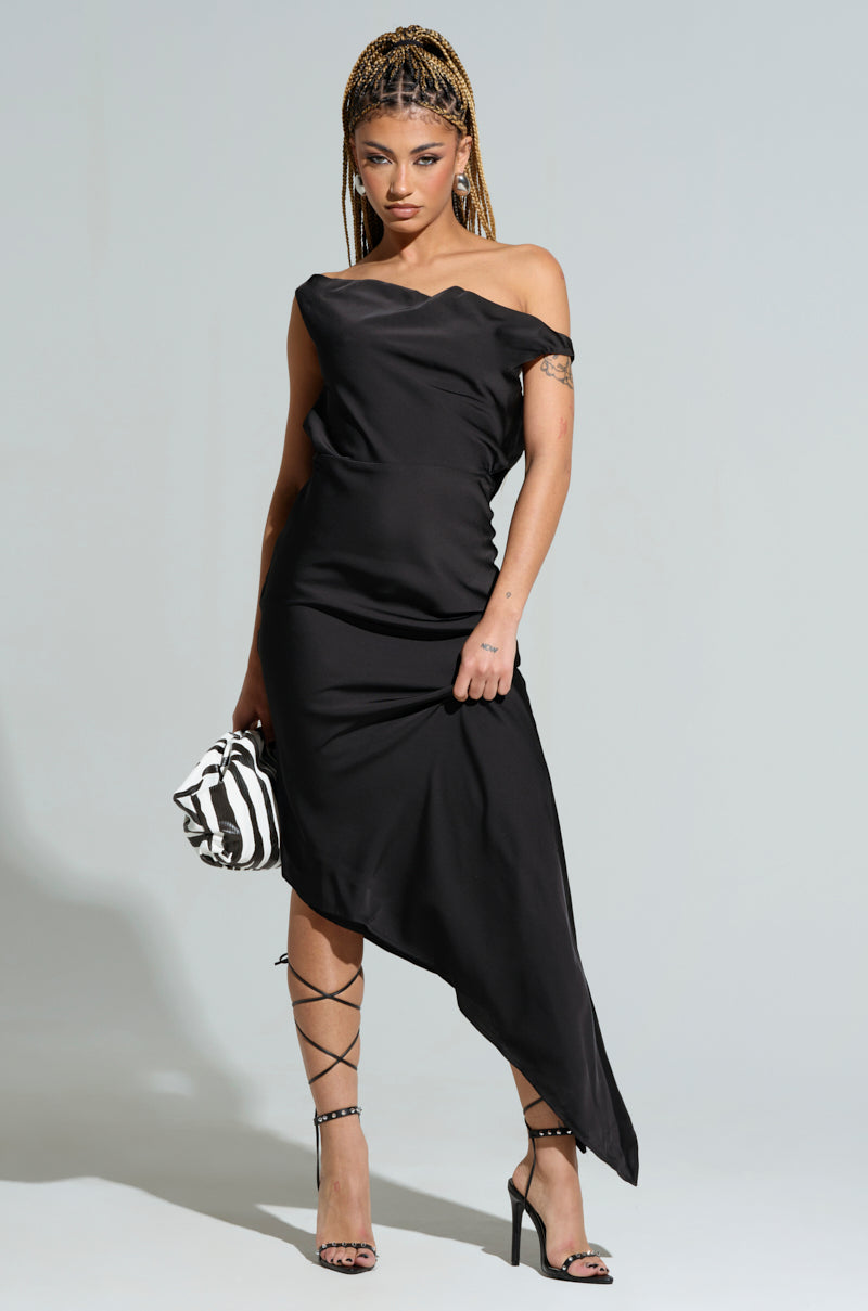 A person poses against a gray background wearing the chic SIMPLY DIVINE SATIN MIDI DRESS accompanied by strappy high heels. Holding a zebra-print handbag, they have braided hair with a tattoo visible on their upper arm. The satin dress showcases an elegant asymmetrical hemline that truly captivates.