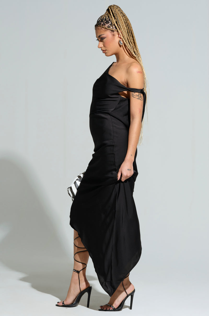 A woman with braided hair is wearing the SIMPLY DIVINE SATIN MIDI DRESS, a sleek black satin midi dress that features an asymmetrical hemline. She complements her look with strappy high heels and carries a small bag. Her arm tattoos are visible as she stands against a plain background.