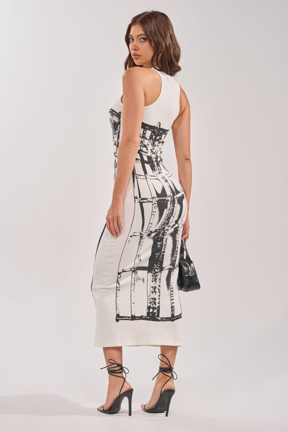 A woman with wavy brown hair models the BREAK FREE PRINTED MIDI DRESS—a sleeveless, racerback white midi with a bold black abstract print. She pairs it with high-heeled black sandals and a small black handbag, glancing over her shoulder.