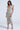 A woman poses confidently in the "I DO WHAT I WANT METALLIC MIDI DRESS IN BEIGE," a sleeveless, metallic midi dress with a plunging V-neckline, front ruching, and silver heels against a light gray background.