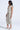 A woman stands in profile wearing the I DO WHAT I WANT METALLIC MIDI DRESS IN BEIGE, featuring a plunging V-neckline and front ruching. She pairs it with silver heels, a silver bracelet, and her long hair is down.