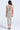 A woman with long dark hair faces away, wearing the I DO WHAT I WANT METALLIC MIDI DRESS IN BEIGE featuring ruched detailing, a low open back tied with a thin string, high heels with ankle ties, and a bracelet.