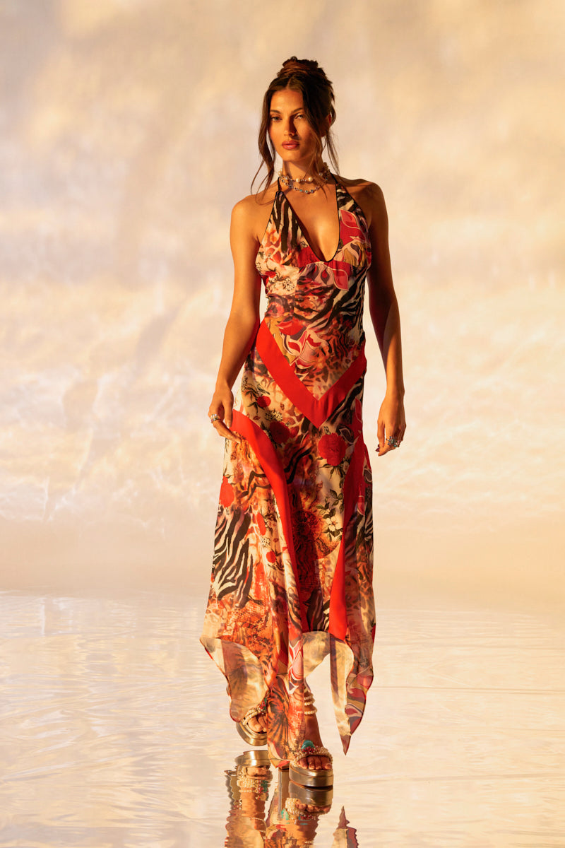 A woman strides confidently on a glossy surface in a vibrant ROTTEN RIGHT TO THE CORE PRINTED MIDI DRESS. Paired with platform sandals, her hair elegantly updo. The softly lit background highlights the warm ambiance of her multi-print ensemble.