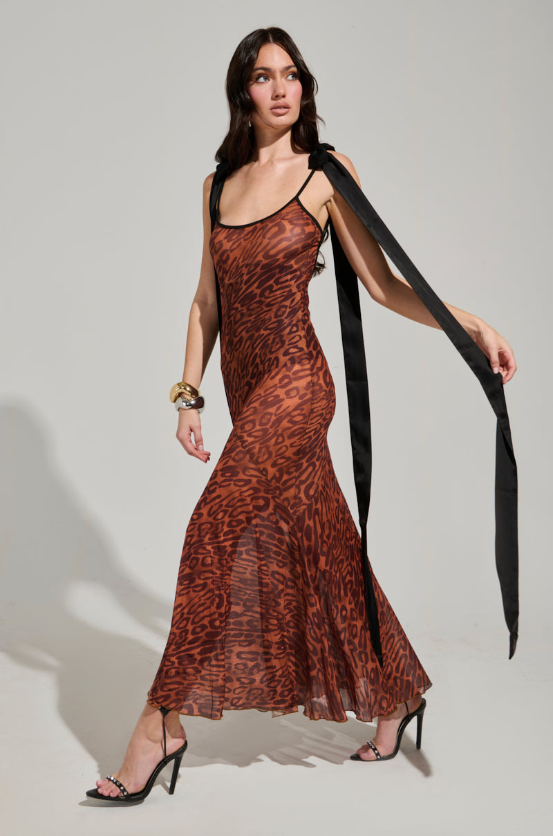 Clad in the ALL ABOUT ME MESH MAXI DRESS, featuring a long and sheer leopard-print design with black straps and flowing ties, an individual strides confidently, complemented by a gold bracelet and black high heels against a light background.