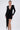A woman models the REBECCA WOVEN MINI DRESS, a fitted long-sleeve mini with padded shoulders, deep V-neckline, and a dramatic draped sash. She styles it with gold earrings, a gold bracelet, and strappy heels against a light background.