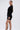 A woman with long straight hair in a ponytail wears the REBECCA WOVEN MINI DRESS—a fitted, long-sleeved black dress with padded shoulders and a tie waist—paired with strappy heels, standing against a white background.