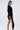 A woman stands in profile against a plain white background wearing the REBECCA WOVEN MINI DRESS—fitted, black, with padded shoulders, long sleeves, and an asymmetrical hem. She accessorizes with gold hoop earrings, a bracelet, and lace-up heels.