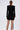 A woman with straight brown hair in a low ponytail models the REBECCA WOVEN MINI DRESS, featuring padded shoulders and a fitted black silhouette, as she stands with her back to the camera against a plain light background.