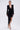 A woman models the REBECCA WOVEN MINI DRESS, a black long-sleeved mini dress featuring padded shoulders, a deep V-neckline, gathered details, and an asymmetric draped panel. She completes the look with gold earrings, a bracelet, and strappy heels.