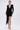A woman stands facing forward against a white background wearing the REBECCA WOVEN MINI DRESS, featuring long sleeves, a deep V-neckline, padded shoulders, and a draped sash. She pairs it with strappy heels, gold jewelry, and has her hair tied back.